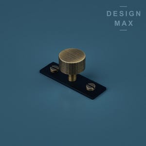 May include: A brass knob with a ridged design, mounted on a black rectangular base with two screws. The knob is a hardware item for furniture or cabinets.