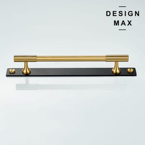 May include: Gold cabinet hardware with a black base plate. The handle is a straight bar with rounded ends and a textured design. The text "DESIGN MAX" is printed above the handle.
