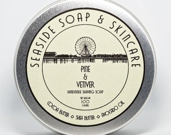 May include: A silver tin with a white label that reads "SEASIDE SOAP & SKINCARE" in a black, stylized font. The label also features a black and white illustration of a Ferris wheel and a pier. The text "PINE & VETIVER HANDMADE SHAVING SOAP" is printed below the illustration. The label also includes the text "NET WEIGHT 100 GRAMS" and "COCOA BUTTER SHEA BUTTER AVOCADO OIL".