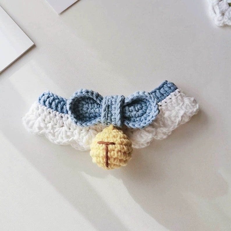 May include: A crocheted pet collar with a blue bow and a yellow bell. The collar is white with a blue crocheted trim.