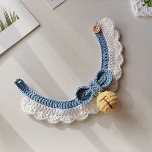 May include: A blue and white crocheted pet collar with a yellow bell and a wooden button. The collar has a scalloped edge and a bow.