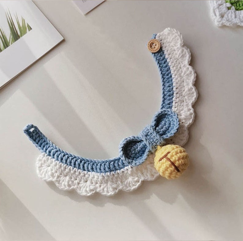May include: A blue and white crocheted pet collar with a yellow bell and a wooden button. The collar has a scalloped edge and a bow.