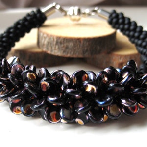 Op de afbeelding: Een zwarte kralen armband met een zilveren sluiting. De kralen zijn in een bloemvormig patroon gerangschikt en hebben een iriserende afwerking.