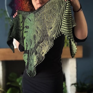 May include: A black and green knitted shawl with a striped pattern. The shawl is held up by a person in front of a fireplace.