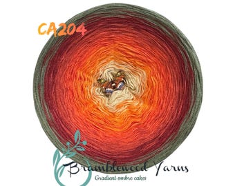 May include: A gradient ombre yarn cake in shades of green, red, orange, and cream. The yarn is wound into a circle with a butterfly design in the center. The label reads "CA204 Bramblewood Yarns Gradient ombre cakes".