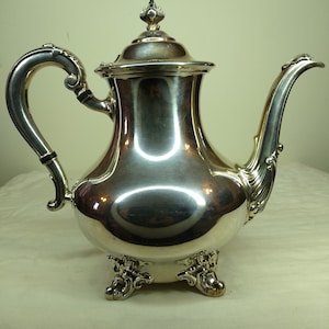 May include: A silver teapot with a rounded body and a decorative handle. The teapot has a lid with a finial and sits on four ornate feet.