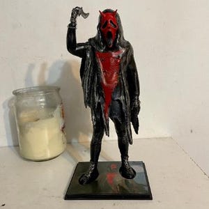 May include: A standing figurine of a character wearing a black hooded robe with a red chest panel and a red mask featuring small horns. The figure holds a small silver-colored blade in its raised right hand and stands on a square black base.