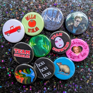 May include: A collection of 11 round button pins with various designs. The pins feature images of a red pickup truck, a tomato, a green background with a person's face, a white egg with a person's face, a rat, a pink background with a heart and a person's face, a black background with a handprint, a black background with text that says "Team Charlie", a green background with text that says "Bella Italia", a blue background with text that says "Team Edward", a black background with text that says "hoa hoa hoa", and a blue background with text that says "titsoak".