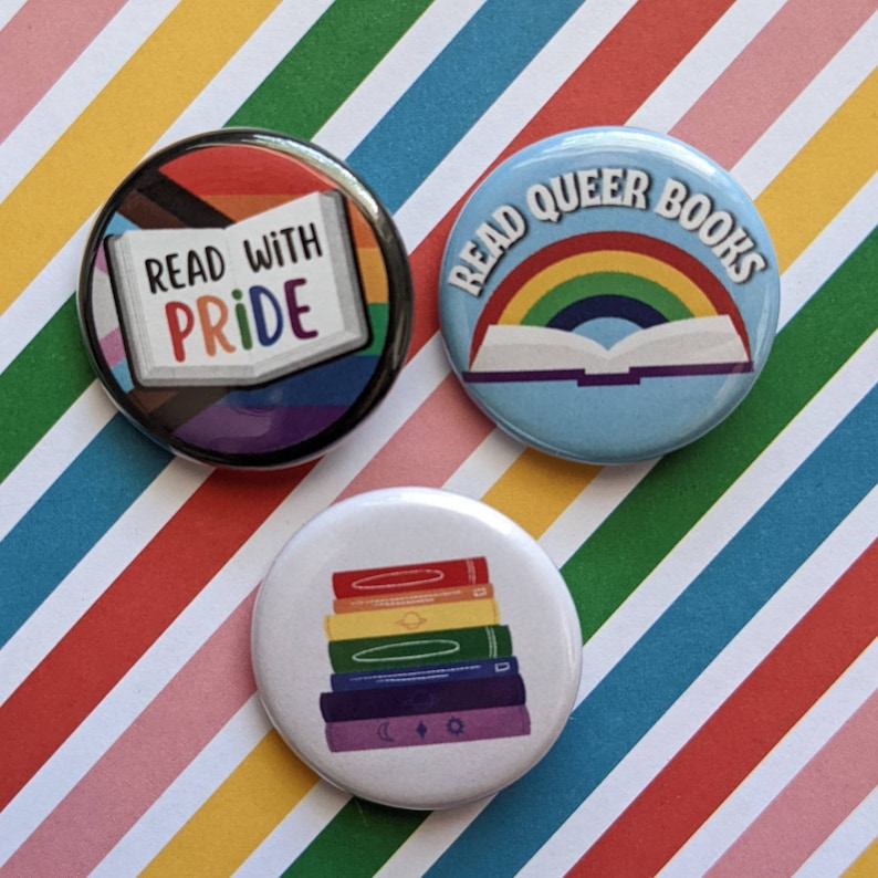 Op de afbeelding: Drie ronde knopen met regenboogontwerpen. De eerste knop zegt "Read with Pride" en heeft een regenboogvlag. De tweede knop zegt "Read Queer Books" en heeft een regenboog en een open boek. De derde knop heeft een stapel regenboogkleurige boeken.