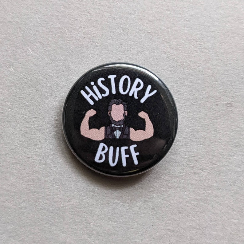 Puede incluir: Un bot&oacute;n negro con texto blanco que dice "HISTORY BUFF" y una imagen de dibujos animados de un hombre con barba flexionando los b&iacute;ceps.