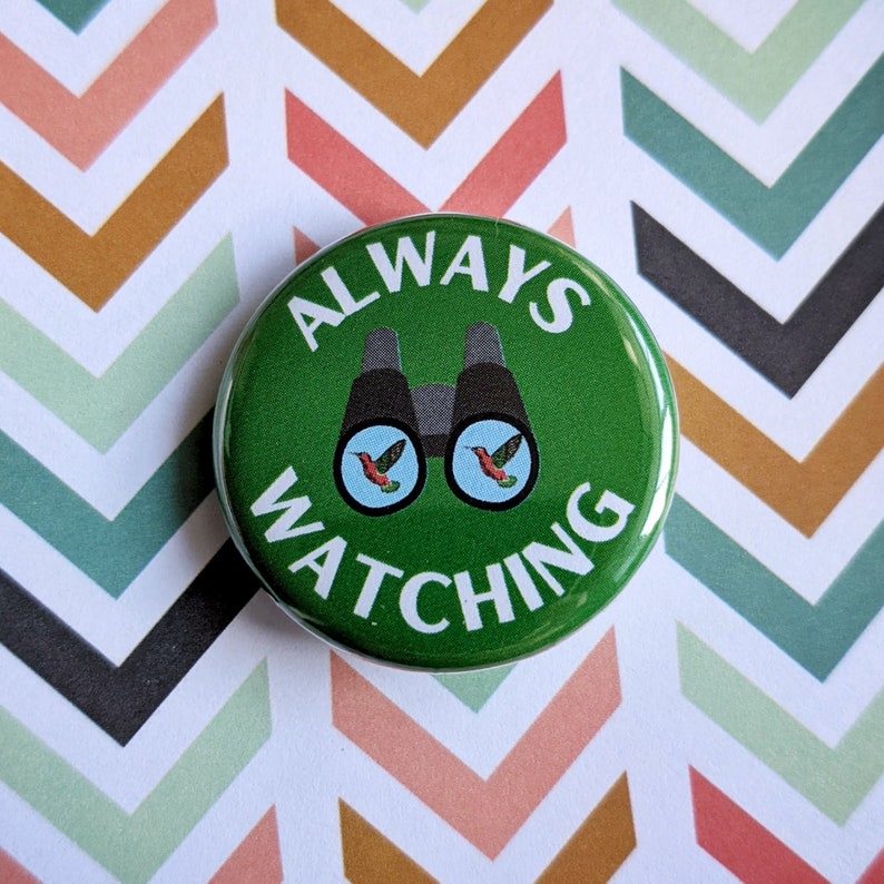 Pode incluir: Um bot&atilde;o verde com o texto "ALWAYS WATCHING" em branco. O bot&atilde;o apresenta um par de bin&oacute;culos pretos com dois beija-flores de peito vermelho olhando atrav&eacute;s das lentes.