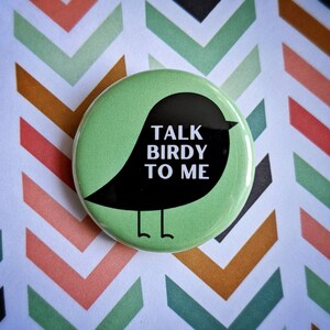 Pode incluir: Um bot&atilde;o verde com uma silhueta preta de um p&aacute;ssaro. O p&aacute;ssaro tem o texto "TALK BIRDY TO ME" escrito nele.