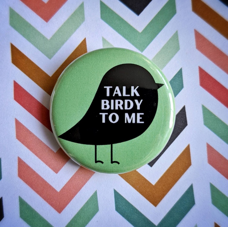 Pode incluir: Um bot&atilde;o verde com uma silhueta preta de um p&aacute;ssaro. O p&aacute;ssaro tem o texto "TALK BIRDY TO ME" escrito nele.