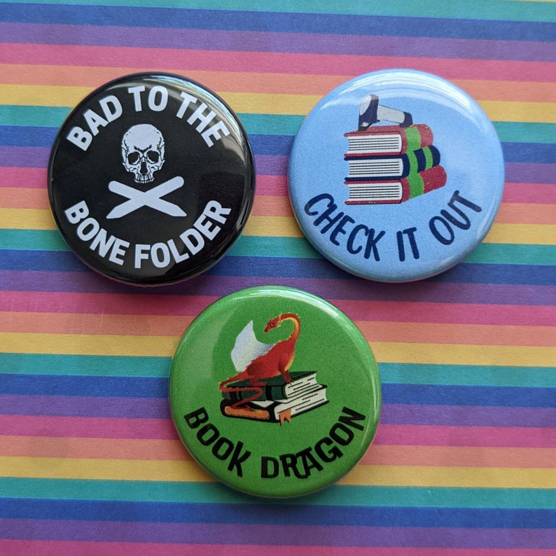 Op de afbeelding: Drie ronde knoppen met verschillende ontwerpen. De eerste knop is zwart met witte tekst die "Bad to the Bone Folder" zegt en een schedel met gekruiste beenderen. De tweede knop is blauw met witte tekst die "Check it Out" zegt en een stapel boeken. De derde knop is groen met witte tekst die "Book Dragon" zegt en een rode draak die op een stapel boeken zit.