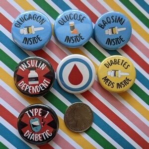 May include: Seven round buttons with various designs related to diabetes. The buttons are blue, yellow, black, and red with white text and images. The text on the buttons includes "Glucagon Inside", "Glucose Inside", "Insulin Dependent", "Diabetes Meds Inside", "Type 1 Diabetic", "Baqsimi Inside", and a red blood drop symbol.