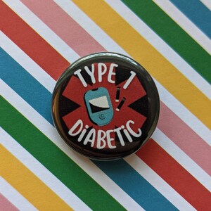 May include: A black and red button with a white star and the text "Type 1 Diabetic" in white. A blue and white medical device and two white insulin needles are inside the star.