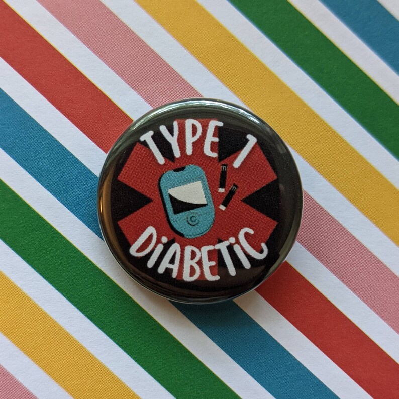 May include: A black and red button with a white star and the text "Type 1 Diabetic" in white. A blue and white medical device and two white insulin needles are inside the star.