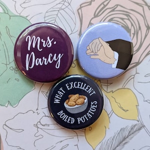 May include: Three round button pins with different designs. The first pin is purple with white text that says "Mrs. Darcy". The second pin is light blue with a cartoon image of two hands holding each other. The third pin is black with white text that says "What Excellent Boiled Potatoes" and a cartoon image of a bowl of potatoes.