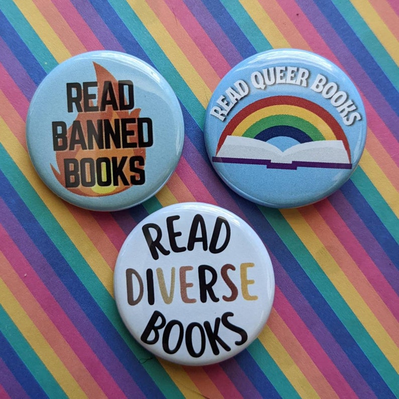 Op de afbeelding: Drie blauwe knoopsbadges met witte tekst en afbeeldingen. De eerste badge zegt "Read Banned Books" met een vlamgrafiek. De tweede badge zegt "Read Queer Books" met een regenboog en een boekgrafiek. De derde badge zegt "Read Diverse Books" met een regenbooggrafiek.