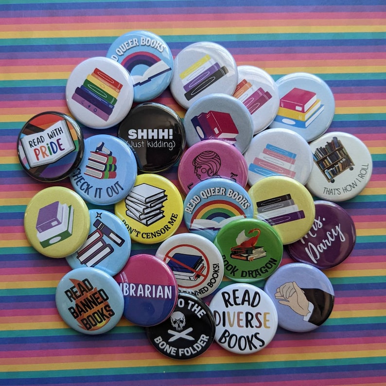 Op de afbeelding: Een verzameling van 18 kleurrijke ronde knoppen met verschillende boekthema's. De knoppen bevatten zinnen zoals "Lees queer boeken", "Lees verboden boeken", "Lees diverse boeken" en "Censureer niet".
