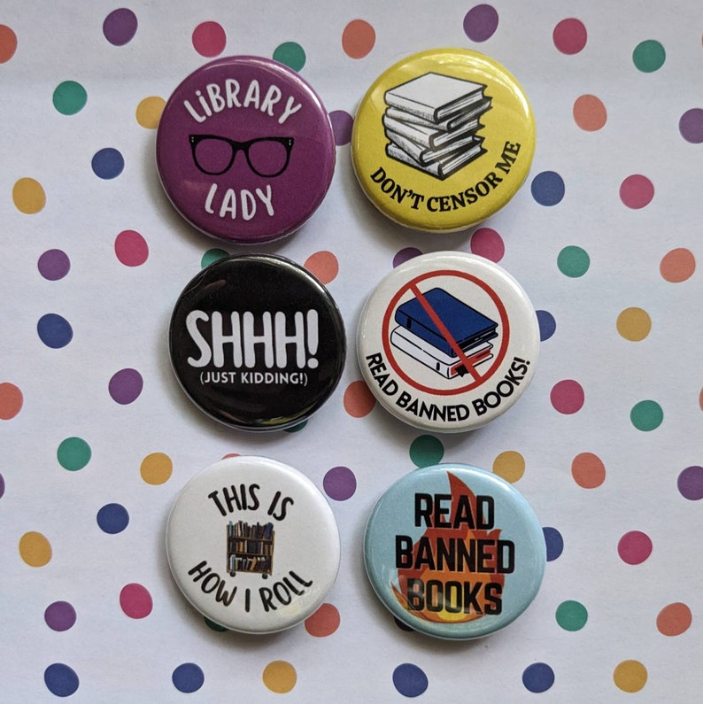 Op de afbeelding: Zes ronde knopen met verschillende ontwerpen met betrekking tot boeken en lezen. De knopen zijn wit, zwart, geel, paars, blauw en rood. De tekst op de knopen is "Library Lady", "Don't Censor Me", "Shhh! (Just Kidding!)", "Read Banned Books", "This is How I Roll" en "Read Banned Books".