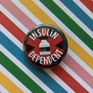 May include: A black and red button with the text "Insulin Dependent" in white lettering. A white vial with the word "insulin" printed on it is in the center of the button.