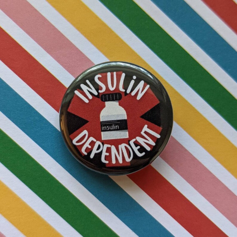 May include: A black and red button with the text "Insulin Dependent" in white lettering. A white vial with the word "insulin" printed on it is in the center of the button.