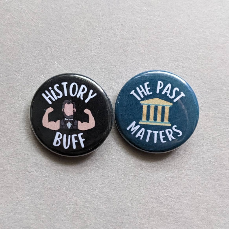 Puede incluir: Dos botones redondos negros y azules con texto blanco. El bot&oacute;n negro tiene una ilustraci&oacute;n de dibujos animados de un hombre con bigote flexionando sus b&iacute;ceps y el texto "HISTORY BUFF". El bot&oacute;n azul tiene una ilustraci&oacute;n blanca de un edificio con columnas y el texto "THE PAST MATTERS".