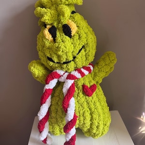 May include: A plush, lime green character with a smiling face, black eyes, and yellow accents. It wears a red and white striped scarf and has a red heart detail. The handmade stuffed animal is displayed on a white surface.