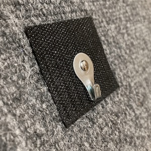 May include: A small, black, square adhesive hook with a silver metal hook. The hook is attached to a gray, fuzzy surface.