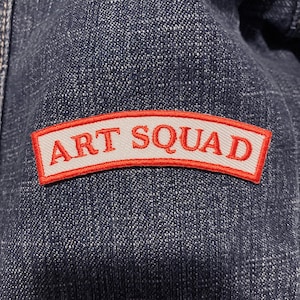 Puede incluir: Primer plano de una tela vaquera azul oscuro con un parche rectangular. El parche tiene un fondo blanco con las palabras "ART SQUAD" en letras rojas, delineadas en rojo. El parche está ligeramente curvado.