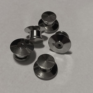 May include: Five silver metal pin backs for attaching badges or other items to clothing. The pin backs have a flat, circular base with a raised center and a small hole in the center.