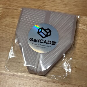 May include: A white 3D printed object in a clear plastic bag. The object has a textured surface and is shaped like a pair of wings. The bag has a sticker with the text "GadCAD3D" and the text "Proudly 3D-Printed in Texas" and the website "www.gadcad3d.com".