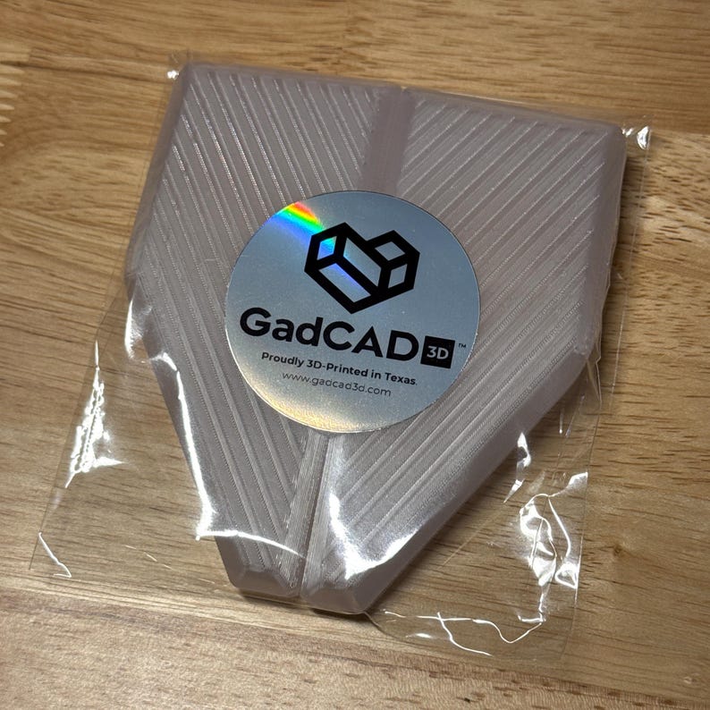 May include: A white 3D printed object in a clear plastic bag. The object has a textured surface and is shaped like a pair of wings. The bag has a sticker with the text "GadCAD3D" and the text "Proudly 3D-Printed in Texas" and the website "www.gadcad3d.com".