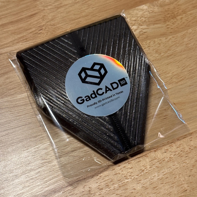 May include: A black 3D printed object with a textured surface. The object is packaged in a clear plastic bag. A silver sticker with the text "GadCAD 3D" and the text "Proudly 3D-Printed in Texas. www.gadcad3d.com" is attached to the object.