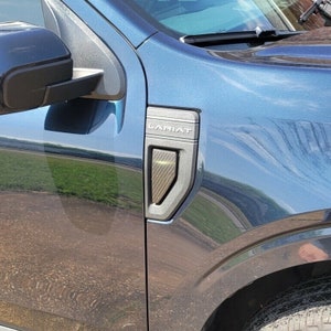 May include: A close-up of a dark blue truck with a black LARIAT emblem on the side. The emblem has a carbon fiber texture and a silver trim.
