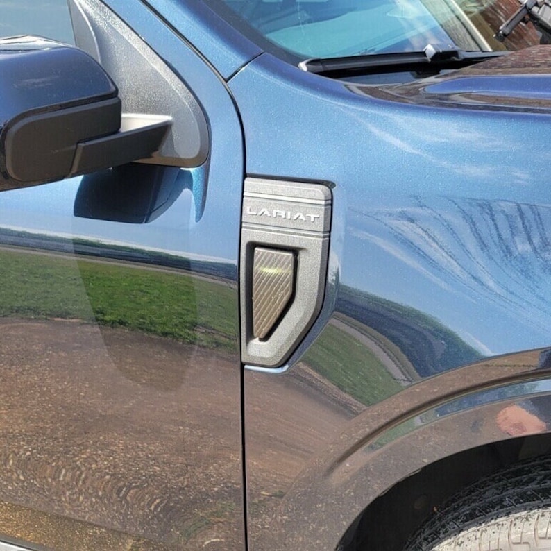 May include: A close-up of a dark blue truck with a black LARIAT emblem on the side. The emblem has a carbon fiber texture and a silver trim.