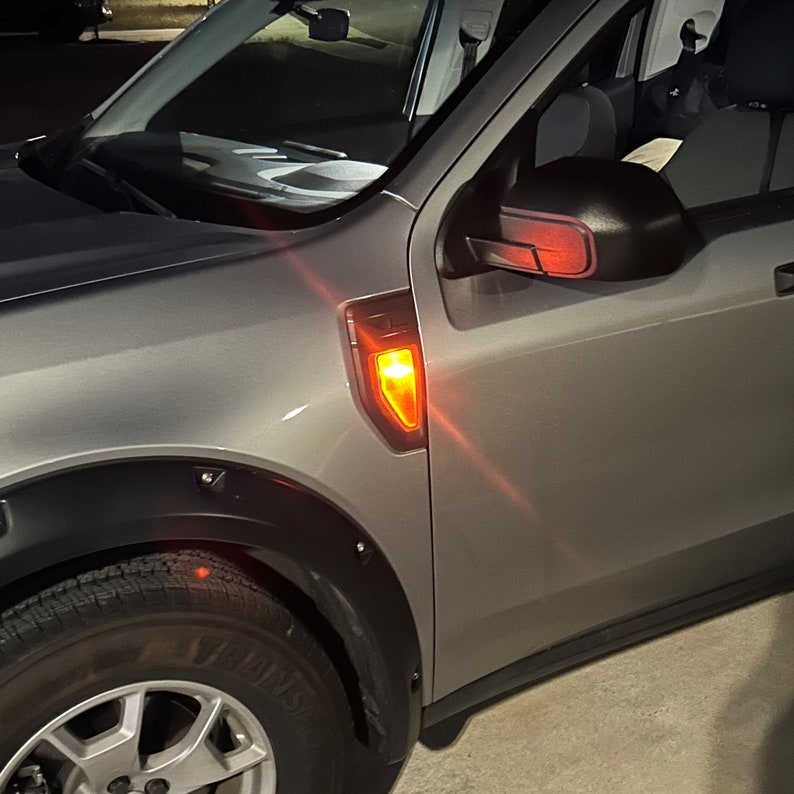 May include: A silver pickup truck with a black fender flare and a red turn signal light. The truck has a black side mirror with a red accent.