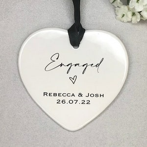 Pode incluir: Adorno de cer&acirc;mica branco em forma de cora&ccedil;&atilde;o com texto preto que diz "Engaged" com um pequeno s&iacute;mbolo de cora&ccedil;&atilde;o e "Rebecca & Josh 26.07.22".