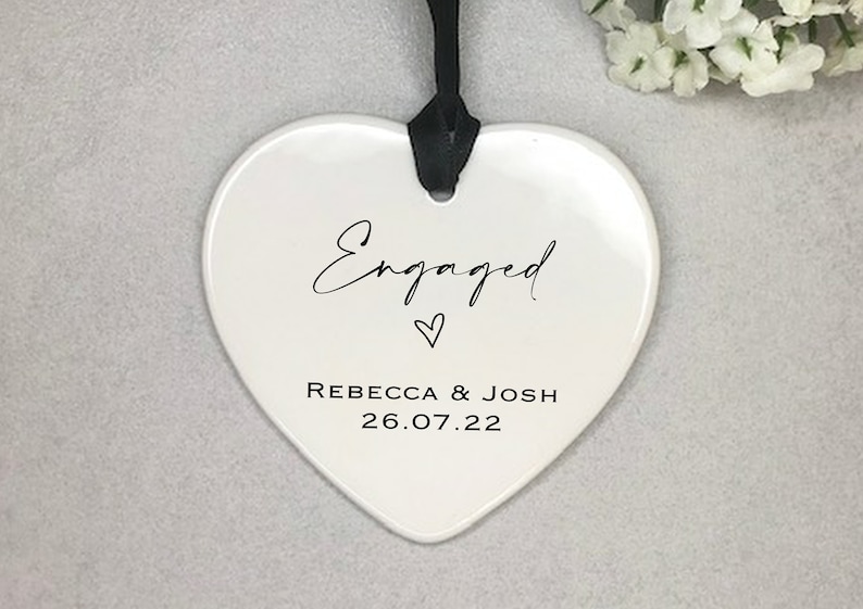 Pode incluir: Adorno de cer&acirc;mica branco em forma de cora&ccedil;&atilde;o com texto preto que diz "Engaged" com um pequeno s&iacute;mbolo de cora&ccedil;&atilde;o e "Rebecca & Josh 26.07.22".