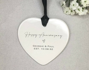 May include: White ceramic heart-shaped ornament with black text that reads "Happy Anniversary" with a small heart symbol, "Georgie & Paul", and "EST. 10.08.92".