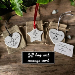 Pode incluir: Tr&ecirc;s ornamentos de cer&acirc;mica branca em forma de estrela e dois cora&ccedil;&otilde;es. O ornamento de estrela tem o texto "Our First Christmas Together" e o texto "Adam & Steve 2024". Os ornamentos de cora&ccedil;&atilde;o t&ecirc;m o texto "Love Congratulations on Earning Your GCSE's Exams" e "Happily Ever After Holly & Tom 30/08/2024". Os ornamentos est&atilde;o em pequenas sacolas de presente de juta com fita vermelha e branca. Um cart&atilde;o branco com o texto "With love on your wedding day Love Mum & Dad" est&aacute; no fundo.