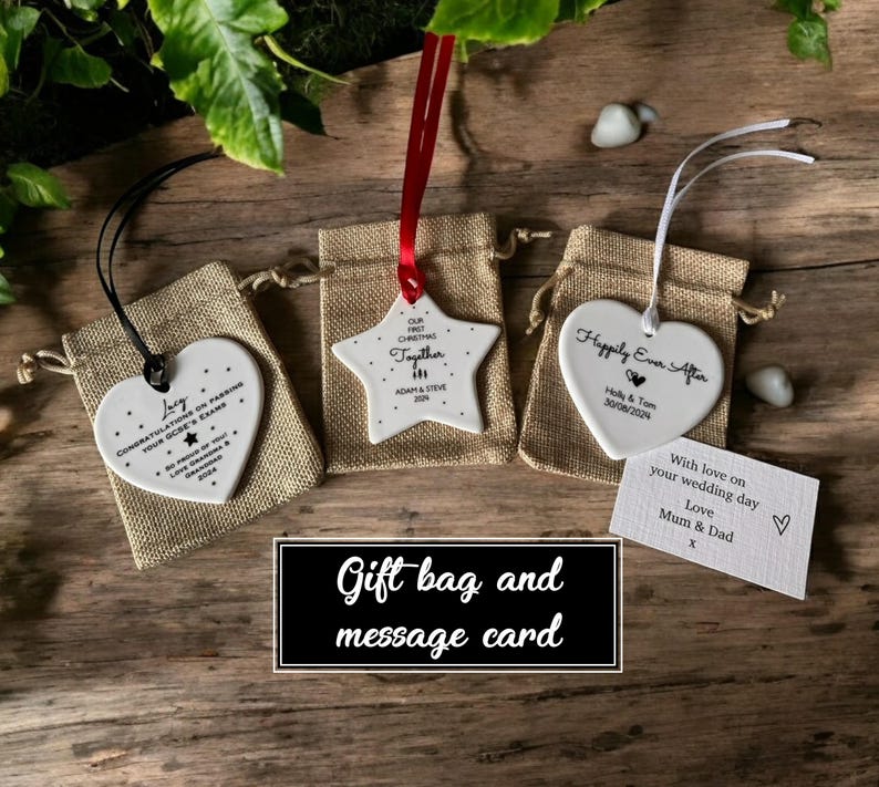 Pode incluir: Tr&ecirc;s ornamentos de cer&acirc;mica branca em forma de estrela e dois cora&ccedil;&otilde;es. O ornamento de estrela tem o texto "Our First Christmas Together" e o texto "Adam & Steve 2024". Os ornamentos de cora&ccedil;&atilde;o t&ecirc;m o texto "Love Congratulations on Earning Your GCSE's Exams" e "Happily Ever After Holly & Tom 30/08/2024". Os ornamentos est&atilde;o em pequenas sacolas de presente de juta com fita vermelha e branca. Um cart&atilde;o branco com o texto "With love on your wedding day Love Mum & Dad" est&aacute; no fundo.