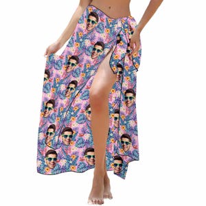 Puede incluir: Un pareo rosa con estampado tropical que presenta un patr&oacute;n repetido de la cara de una persona con gafas de sol. El pareo est&aacute; atado a la cintura y tiene una abertura en el lateral.
