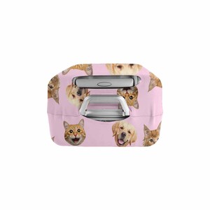 May include: A pink suitcase cover with a repeating pattern of cat and dog faces. The cover has cutouts for the handle and is designed to fit over a suitcase. The animal faces are in shades of orange, brown, and gold.