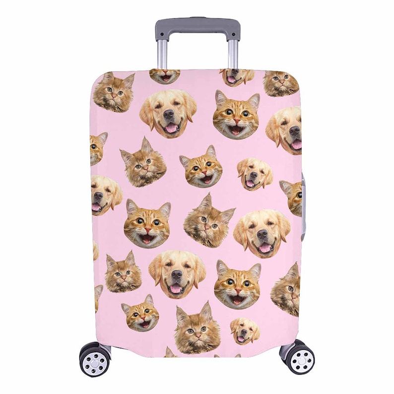 May include: A pink suitcase cover with a repeating pattern of smiling golden retriever and ginger cat faces. The luggage cover has a retractable handle and two wheels.