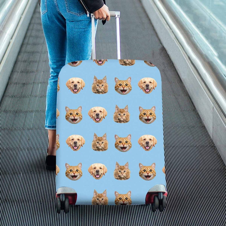 May include: A light blue suitcase with a repeating pattern of smiling golden retriever and cat faces. The suitcase has a silver handle and black wheels. The person holding the suitcase is wearing blue jeans.