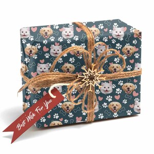 May include: A blue gift wrap with a repeating pattern of dog and cat faces, pink hearts, and white paw prints. The gift is wrapped with twine and a wooden snowflake ornament. A red gift tag with the text "Best Wish For You" is attached to the gift.