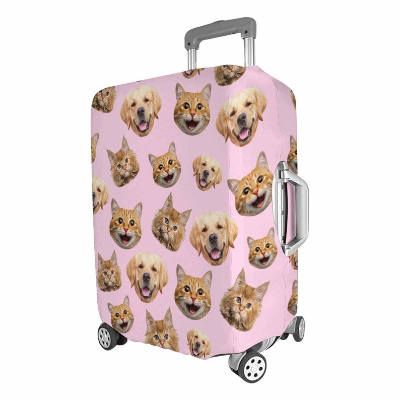 May include: A pink suitcase cover with a repeating pattern of smiling golden retriever and ginger cat faces. The suitcase has a retractable handle and wheels, designed for travel. The cover is made of a durable material.