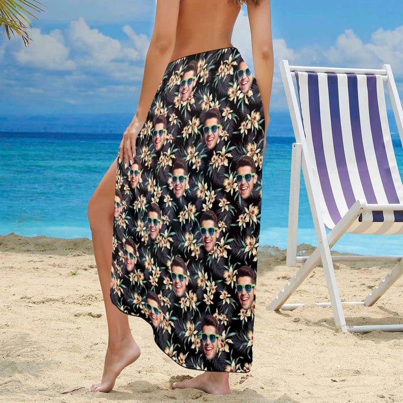 Puede incluir: Un sarong negro con un patr&oacute;n floral que presenta una imagen repetida de una persona con gafas de sol. El sarong lo lleva una persona de pie en una playa de arena.